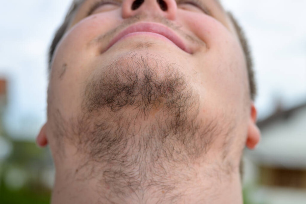 Joven sin afeitar con una barba enmarañada en primer plano en su garganta y el pelo facial al aire libre contra el cielo gris - Foto, imagen