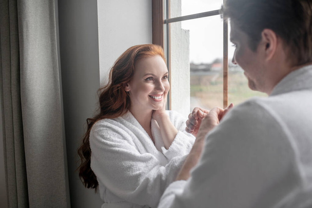 A couple in white bathrobes standing near the window and looking romantic - Fotografie, Obrázek