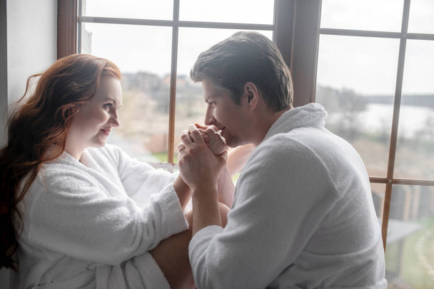 A couple in white bathrobes standing near the window and looking romantic - Fotoğraf, Görsel