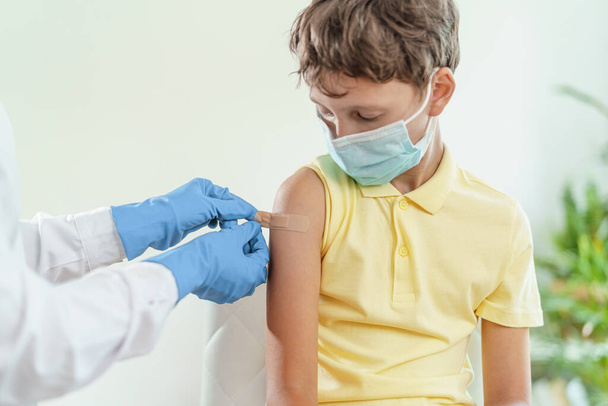 el médico le administra a un niño con una máscara médica una inyección en el hombro. Vacunación de niños contra el coronavirus. La vacuna contra Covid-19 - Foto, Imagen