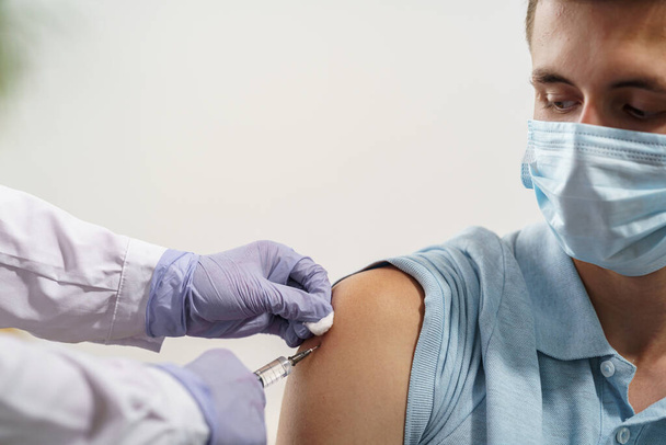 doctor gives an adult young man in a medical mask an injection in the shoulder. Vaccination of adults against coronavirus. vaccine against Covid-19. Protection against the virus. Close up. copy space - Photo, Image