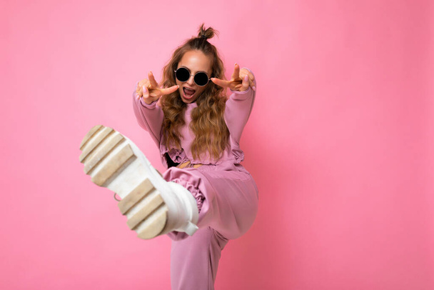 Photo of happy positive young attractive blonde curly woman with sincere emotions wearing trendy pink sportswear and sunglasses isolated on pink background with copy space and having fun - Photo, Image