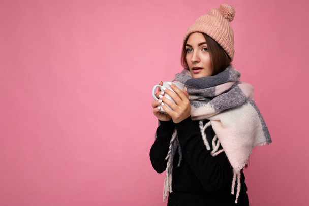 美しい若いですブルネット女性身に着けている黒セーター帽子と暖かいスカーフ隔離された上ピンクの背景保持紙マグカップ用mokup飲料熱いお茶と見てカメラと持っている寒さ - 写真・画像