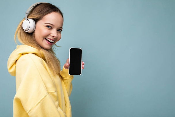 Boczny profil zdjęcie pięknej szczęśliwej uśmiechniętej młodej kobiety noszącej stylowy strój casual izolowane na tle ściany gospodarstwa i pokazując telefon komórkowy z pustym wyświetlaczem do makiety noszenia biały - Zdjęcie, obraz