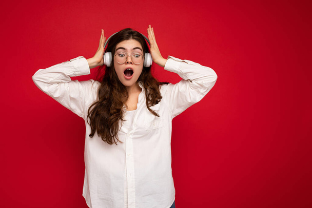 Приваблива шокована молода брюнетка жінка в білій сорочці та оптичних окулярах ізольована на червоному тлі в білих бездротових навушниках Bluetooth, що слухають музику та дивляться вниз
 - Фото, зображення