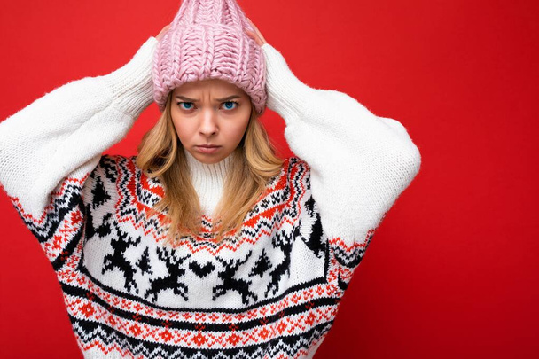 Photo of beautiful dissatisfied sad young blonde woman isolated over red background wall wearing winter sweater and pink hat looking at camera - 写真・画像