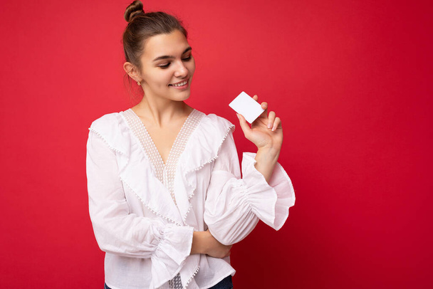 Hermosa feliz sonriente joven rubia oscura con blusa blanca aislada sobre fondo rojo sosteniendo tarjeta de crédito mirando a la tarjeta de plástico - Foto, Imagen
