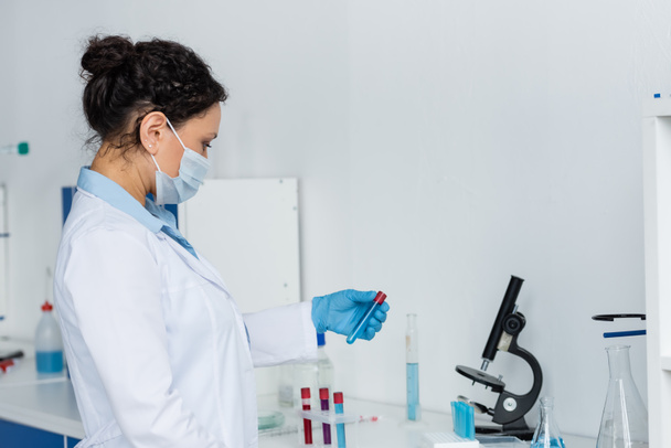 Tıbbi maskeli ve beyaz önlüklü Afro-Amerikan bilim adamının laboratuardaki deney tüplerinin yanında deney tüpü tutuşu.  - Fotoğraf, Görsel