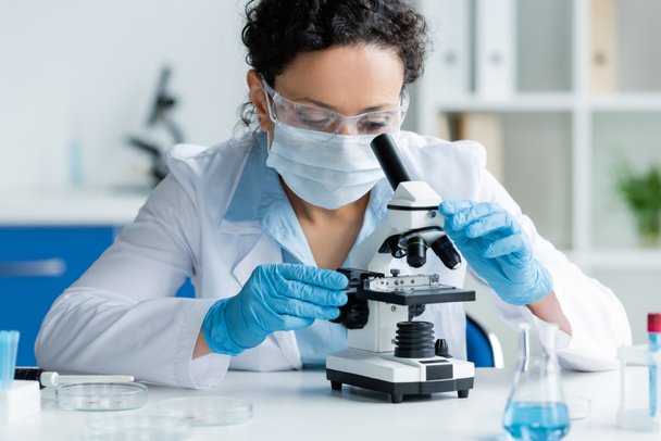 Cientista afro-americano em máscara médica e luvas de látex olhando através de microscópio perto de placas de Petri  - Foto, Imagem