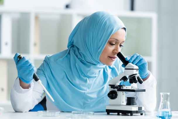 Arabischer Wissenschaftler im Hijab schaut durch das Mikroskop und hält eine elektronische Pipette  - Foto, Bild