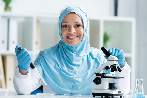 uśmiechnięty arabski naukowiec trzymający pipetę i mikroskop podczas pracy w laboratorium  - Zdjęcie, obraz