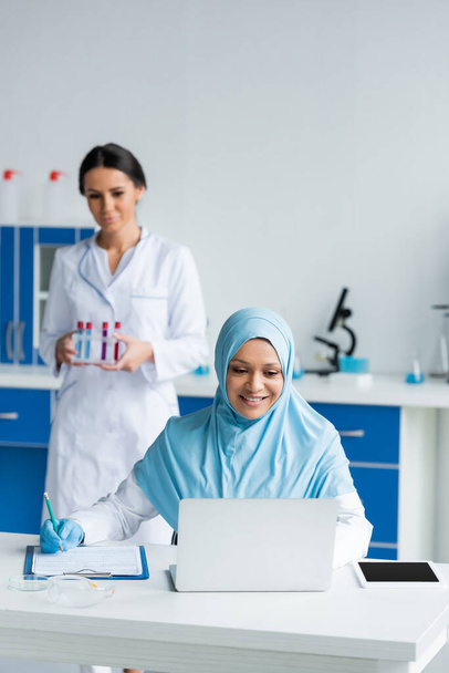 Smiling muslim scientist using laptop and writing on clipboard near blurred colleague  - Foto, Imagen