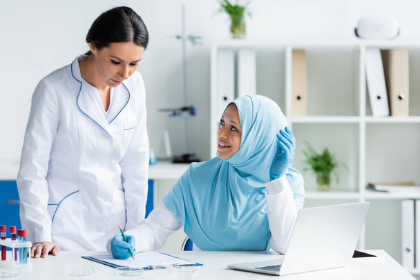 Cheerful muslim scientist in latex gloves writing on clipboard and looking at colleague near laptop in laboratory  - Photo, Image