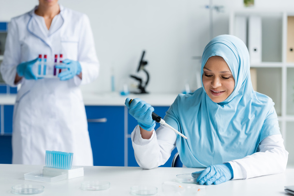 Un scientifique arabe souriant tenant une pipette électronique près de boîtes de Pétri et un collègue  - Photo, image