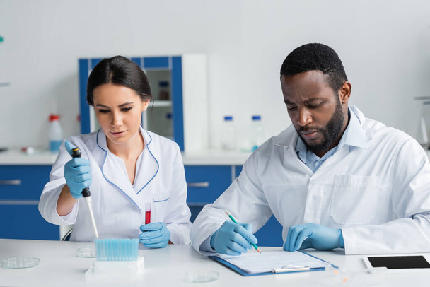 African american scientist writing on clipboard near colleague with pipette and test tubes  - Foto, Imagen