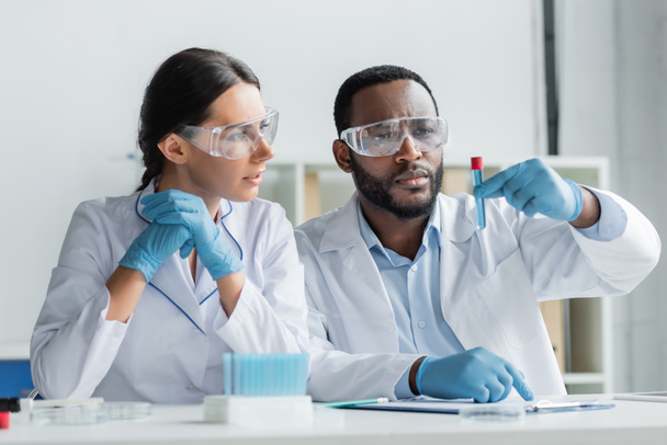 African american scientist in safety goggles holding test tube near colleague in latex gloves  - Foto, Imagen