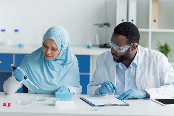 Afrykańscy amerykańscy i arabscy naukowcy pracujący z płytami Petriego, tabletem cyfrowym i schowkiem w laboratorium  - Zdjęcie, obraz