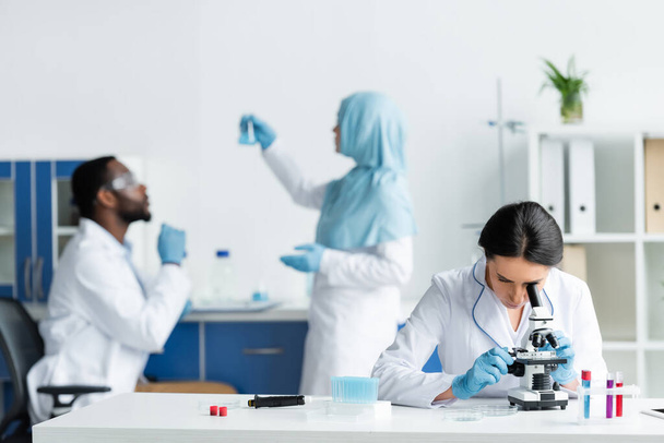 Scientist looking through microscope near blurred interracial colleagues in lab - Foto, Imagen