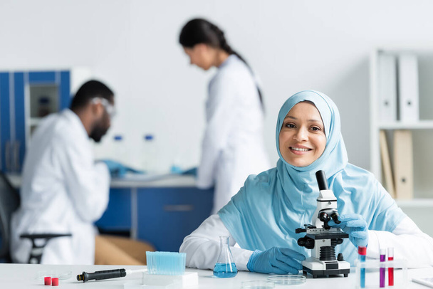 Scientifique musulman positif regardant la caméra près du microscope, la fiole et la pipette en laboratoire  - Photo, image