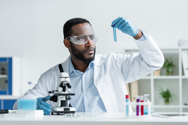 African american scientist in latex gloves and safety goggles holding test tube near microscope  - Foto, Imagen