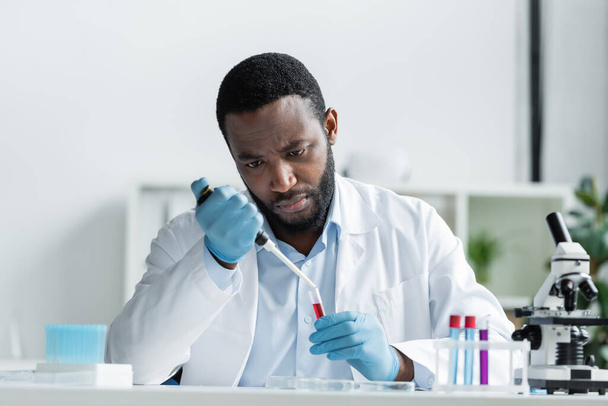 African american scientist in latex gloves holding test tube and electronic pipette in laboratory  - Foto, Imagen