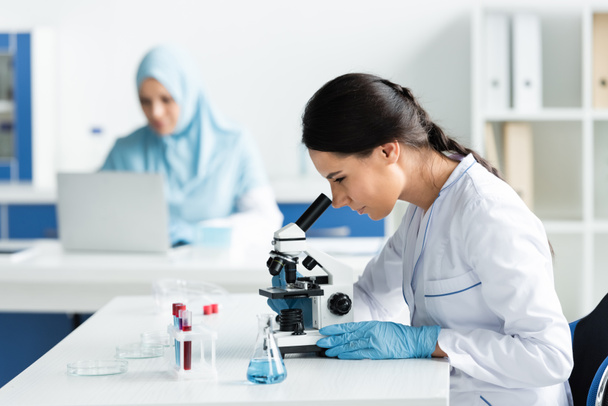Науковець у латексних рукавичках дивиться через мікроскоп поблизу колби з реагентом і тестовими трубками  - Фото, зображення