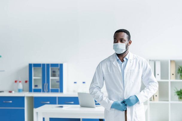 African american scientist in medical mask adjusting white coat in lab  - Photo, Image