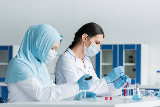 Side view of scientist in protective mask working near arabian colleague with pipette on blurred foreground  - 写真・画像