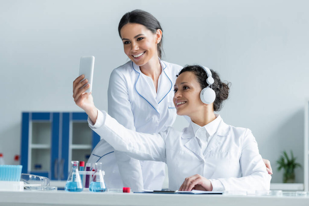 Cientista afro-americano sorridente em fones de ouvido usando smartphone perto colega em laboratório  - Foto, Imagem
