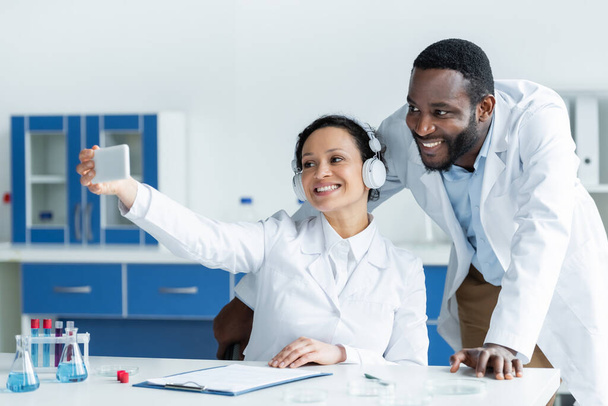 Un scientifique afro-américain dans un casque utilisant un smartphone près d'un collègue souriant dans un laboratoire  - Photo, image