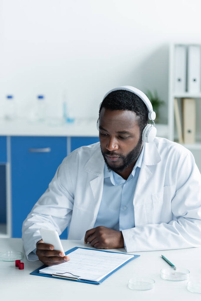African american scientist in headphones using smartphone near petri dishes and clipboard  - Фото, изображение