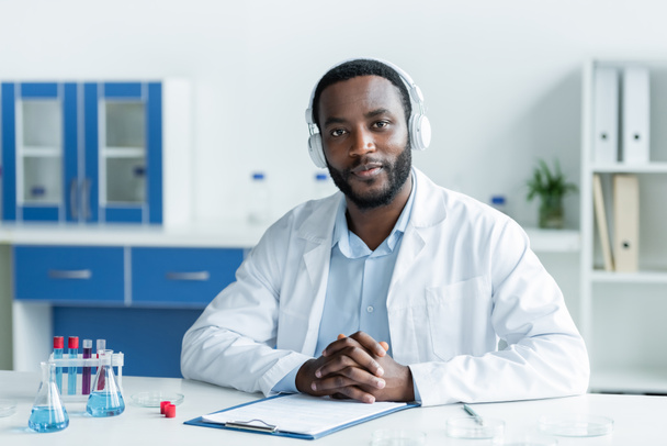 African american scientist in headphones looking at camera near flasks and clipboard  - Photo, Image
