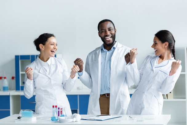 Excited interracial scientists showing yes gesture near smartphone, clipboard and test tubes  - Photo, Image