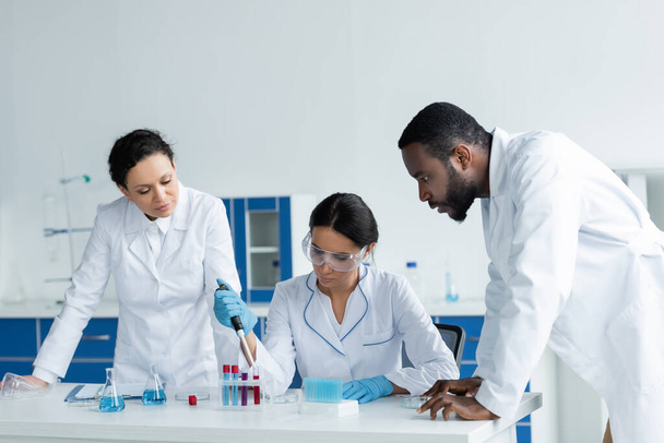 Scientist working with pipette and test tubes near focused colleagues in lab  - Photo, Image