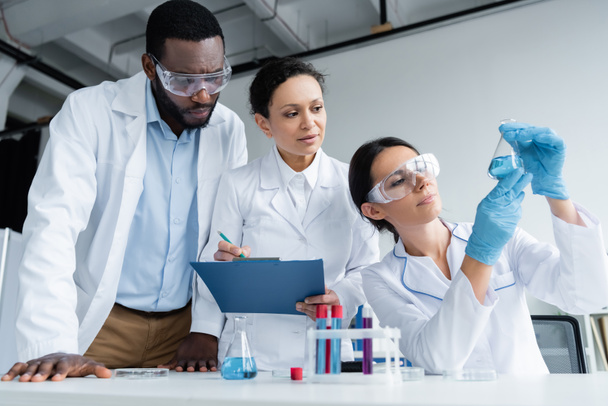 Interracial scientists in safety goggles with clipboard looking at reagent in flask  - Foto, Imagen