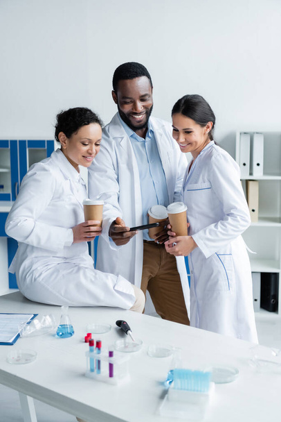 Positive interracial scientists with coffee looking at smartphone in laboratory  - Photo, Image