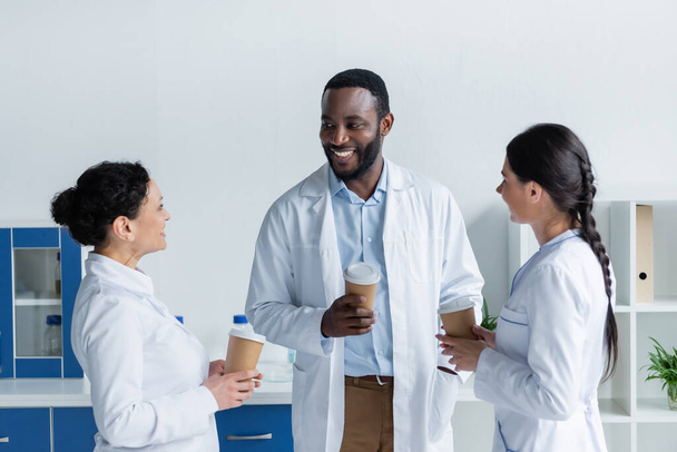 счастливые многонациональные врачи, держащие бумажные стаканчики в клинике  - Фото, изображение