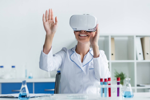 Smiling scientist in vr headset sitting near test tubes on blurred foreground  - Photo, Image
