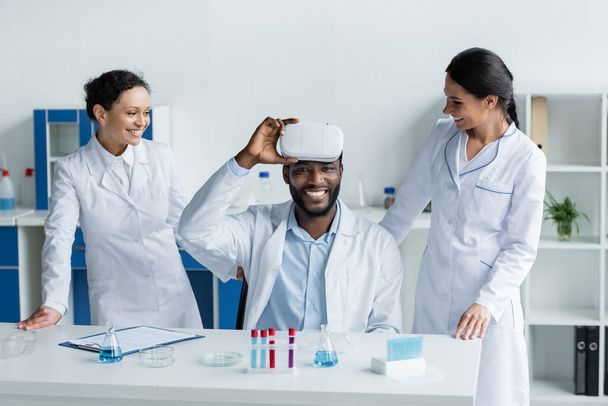 Cheerful african american scientist holding vr headset near colleagues in laboratory  - Photo, Image
