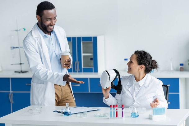 Un scientifique afro-américain avec du café pour pointer du doigt un collègue avec un casque vr en laboratoire  - Photo, image