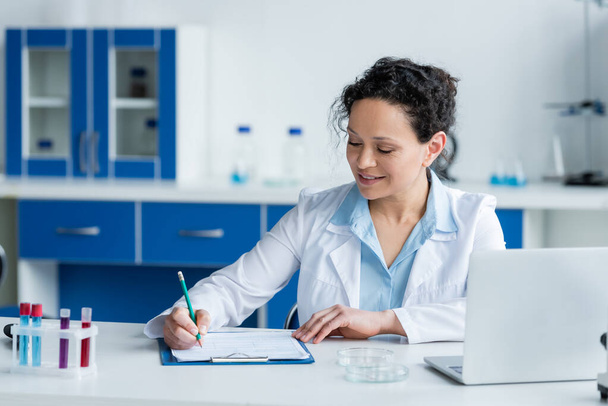Smiling african american scientist writing on clipboard near laptop and petri dishes  - Фото, изображение