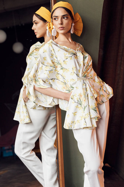 photo of a girl near a full-length mirror looking in the direction of wearing a white blouse with yellow flowers and white pants looking to the side and wearing a yellow headband - Foto, imagen