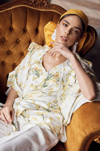 portrait photo of a girl with brown eyes with a yellow bow on her head in a blouse with yellow flowers and white pants sitting on the couch - Valokuva, kuva