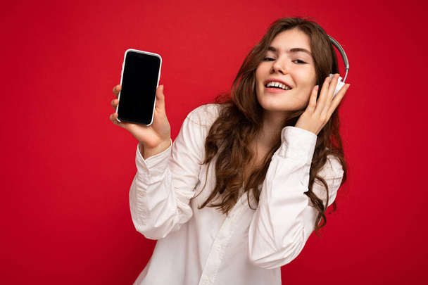 Hermosa mujer joven sonriente feliz con elegante atuendo casual aislado en la pared de fondo sosteniendo y mostrando el teléfono móvil con pantalla vacía para maqueta usando auriculares bluetooth blancos - Foto, imagen