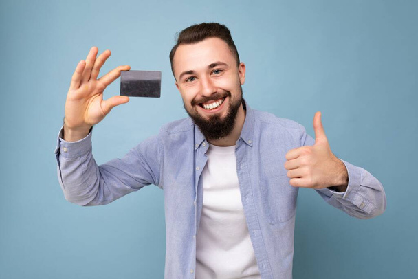 Closeup portrait of Good looking cool smiling brunette bearded young man wearing stylish blue shirt and white t-shirt isolated over blue background wall holding credit card looking at camera and - Photo, Image
