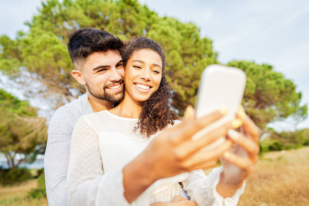Młoda, piękna, wielorasowa para zakochana w przyrodzie robi sobie selfie ciesząc się wakacyjnymi chwilami, dzieląc się zdjęciami w sieciach społecznościowych. Nowe normalne uzależnienie od technologii dzięki bezprzewodowym połączeniom internetowym Wi-Fi - Zdjęcie, obraz