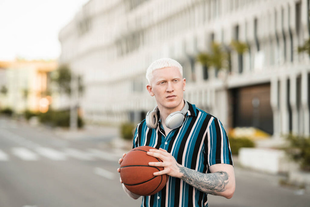 Junger, nachdenklicher Albino-Blondschopf mit Basketballball und Kopfhörer, der in der Stadt spaziert - Foto, Bild
