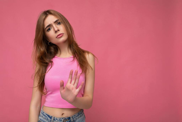 Serious upset young attractive dark blonde woman with sincere emotions isolated on background wall with copy space wearing stylish pink top and doing stop gesture. Negative concept - Foto, afbeelding