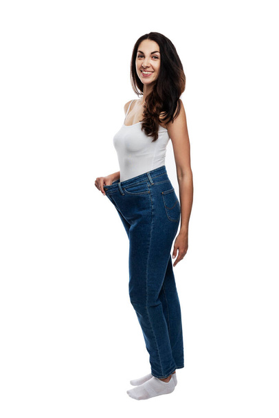 Young woman in jeans one size up. Happy beautiful brunette. Healthy lifestyle and slimness. Full height. Isolated on white background. Vertical. - Foto, Imagen