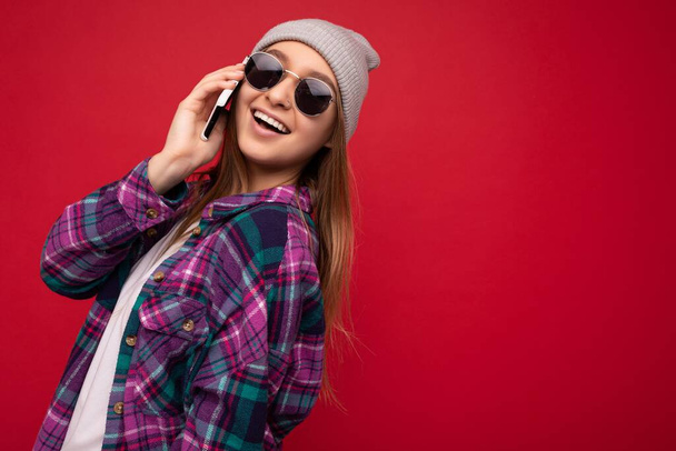 foto de hermosa mujer rubia joven positiva feliz vistiendo camisa púrpura hipster y casual camiseta blanca sombrero gris y gafas de sol aislados sobre fondo rojo sosteniendo en la mano y hablando en el móvil - Foto, imagen